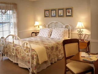 A cozy bedroom with a floral-patterned bed, two bedside lamps, a window with lace curtains, four framed pictures on the wall, and a small writing desk with a chair.