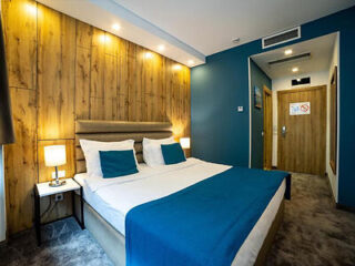 A cozy hotel room with a double bed, wooden accent wall, blue decor, and two bedside tables with lamps. A hallway leads to the entrance door, which has a no-smoking sign.