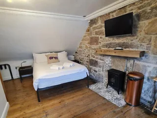 A cozy bedroom with a stone accent wall, a small bed, a wall-mounted TV, folded towels, and a wood-burning stove. The room has wooden flooring and minimal furniture.