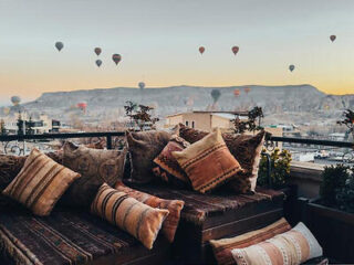 A rooftop terrace with cushioned seating overlooks a landscape where numerous hot air balloons float in the sky at dawn.