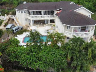 An aerial view of a large white house with a pool in the backyard, surrounded by greenery. Two parked cars are visible near the building.