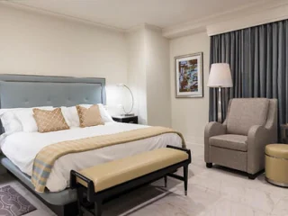 A neatly made bed with beige and white bedding, a bench at the foot, an armchair, a side table with a lamp, and a curtained window in a modern bedroom.