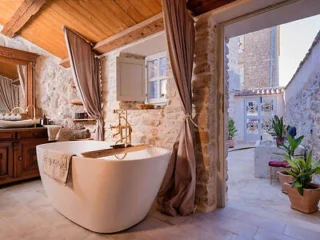 Rustic bathroom with stone walls, a freestanding tub with a towel, wooden ceiling, and vanity. Curtains frame an open doorway leading to a sunlit outdoor area with plants and a small seating space.