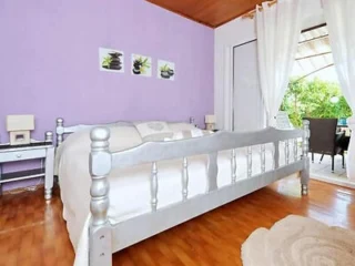 A bedroom with a purple wall, a large bed with light-colored bedding, two side tables with lamps, and framed artwork above the bed. A sliding door leads to an outdoor area with seating and greenery.