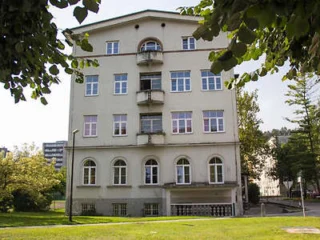 A four-story beige apartment building with multiple windows and small balconies, surrounded by trees and grass.