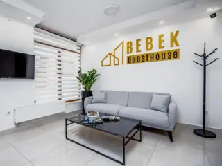 A bright and welcoming guesthouse lounge with a light gray sofa, a modern coffee table, and a sign reading "Bebek Guesthouse" on the wall.