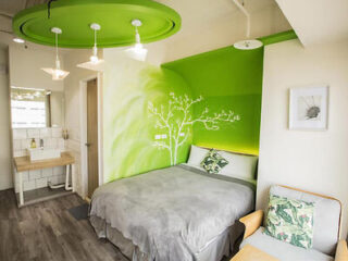 A brightly lit bedroom with green and white walls, a round ceiling accent, a bed with gray bedding, and leafy pillows. A sink is seen in the corner next to a door and a wall-mounted painting.