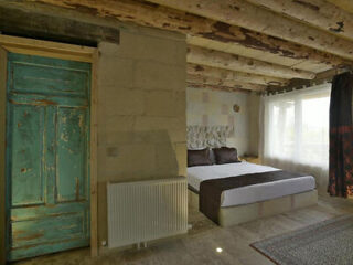 A rustic bedroom with a large bed, textured walls, wooden ceiling beams, and a green wooden door. Sunlight brightens the room through a window with sheer and dark curtains.