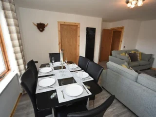 A dining room with a black table set for six, adjacent to a living area with a gray couch. The room features wooden doors, a window with plaid curtains, and a mounted wall decoration.