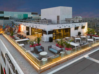 A rooftop bar and lounge area with modern outdoor furniture, string lights, plants, and wall murals, set against a backdrop of city buildings during evening time.