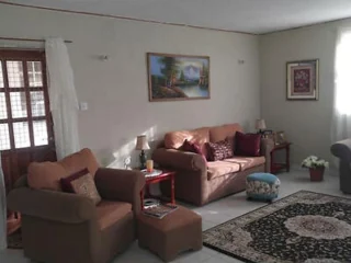 A living room with two brown couches, a small cushioned ottoman, a rug, framed artworks on the walls, and a side table with a lamp. White curtains cover the glass door to the left.
