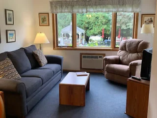 A living room with a gray couch, a brown recliner, a wooden coffee table, a TV on a stand, two lamps, framed pictures on the walls, and a large window showing an outdoor scene with a gazebo.