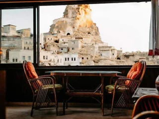 Two wicker chairs with orange cushions and a table are set in front of a large window displaying a view of a historic stone structure built into a rocky hillside.
