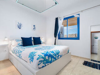 A bedroom with a blue and white ocean-themed decor, featuring a bed with coral-patterned bedding, two navy blue pillows, white walls adorned with sea life artwork, a window with blue trim, and tiled floor.