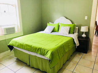 A bedroom with a green theme, featuring a double bed with green bedding and pillows, a bedside table with a lamp, a window with an air conditioning unit, and tiled flooring.