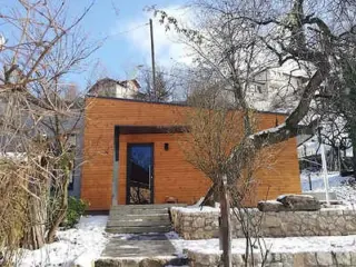 A charming wooden house surrounded by a winter garden, with a path leading to the entrance, set against a snowy backdrop.
