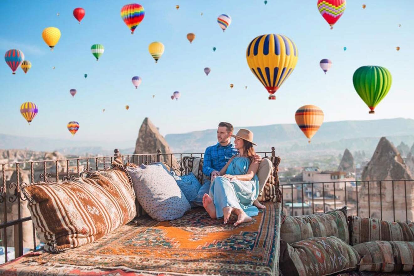 A couple sits on a cushioned outdoor terrace overlooking a landscape filled with colorful hot air balloons floating in the sky.