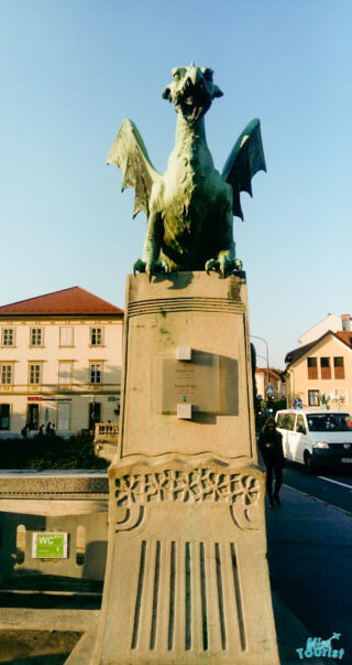 A stone statue of a dragon on a pedestal stands against a backdrop of buildings and a clear sky. An information plaque is affixed to the pedestal.