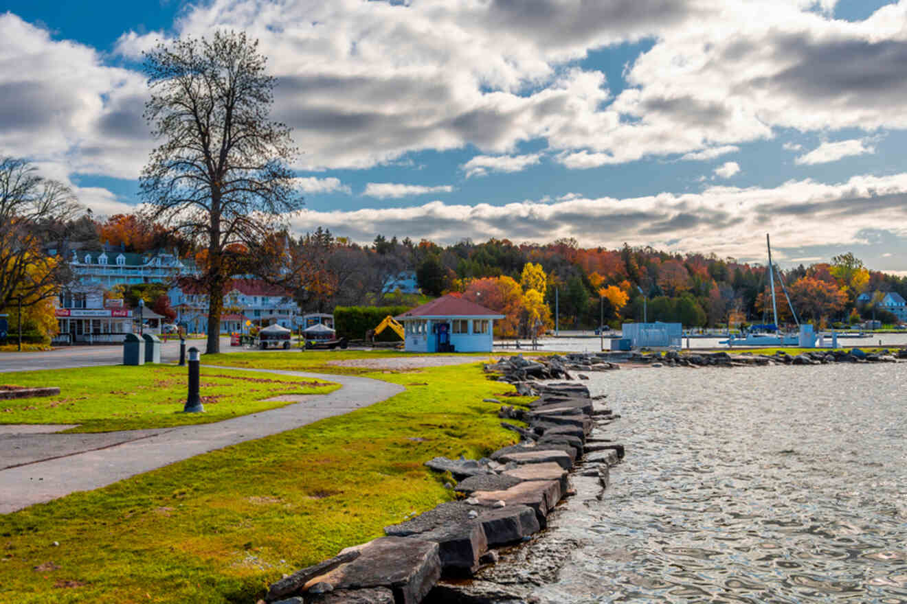 A waterfront park with a rocky shoreline, a path, and buildings surrounded by fall foliage under a partly cloudy sky.