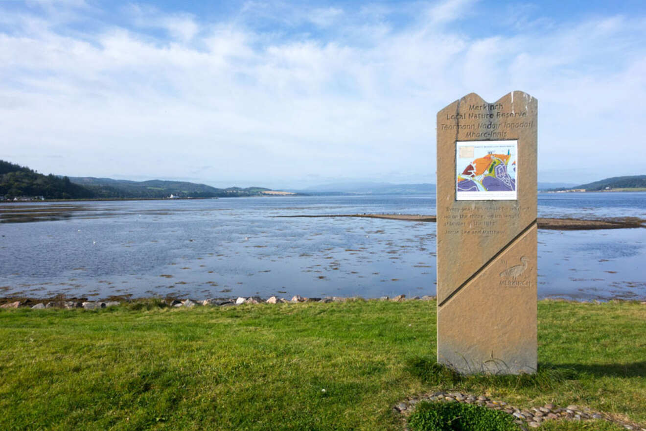 A standing stone slab with colorful artwork and text stands on a grassy area overlooking a calm bay and distant hills under a clear sky.
