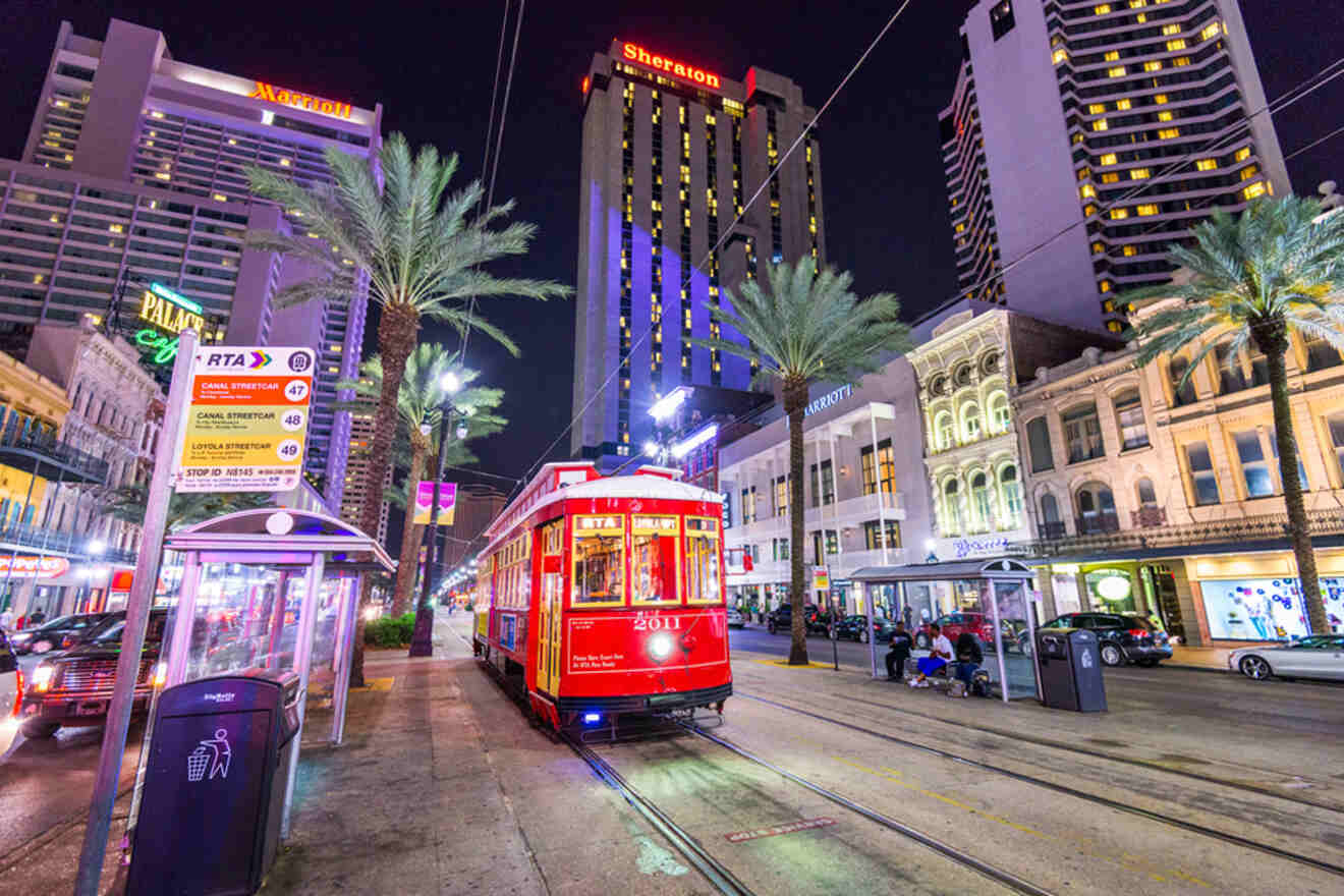 A red streetcar passes through a brightly lit downtown street at night, surrounded by tall buildings, palm trees, and street signs.