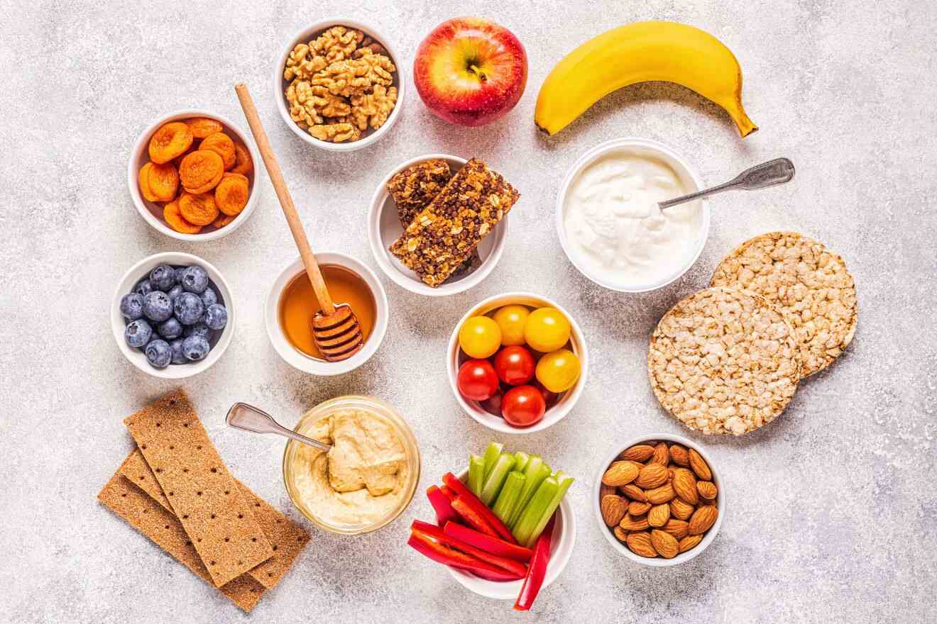 A variety of snacks including dried apricots, walnuts, an apple, a banana, blueberries, honey, granola bars, yogurt, rice cakes, cherry tomatoes, hummus, celery, bell peppers, crackers, and almonds.