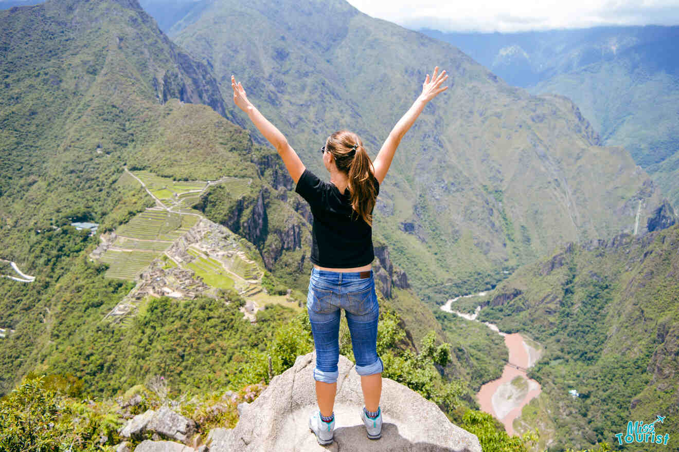 Yulia, la fundadora de este sitio web, se encuentra en el borde de una roca, con los brazos en alto y la espalda de espaldas a la cámara, contemplando un vasto paisaje montañoso. Al fondo se puede ver Machu Picchu, entre los verdes picos.