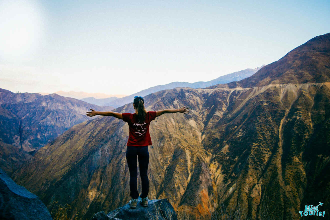 Yulia, la fundadora de este sitio web, se encuentra de pie sobre una roca con los brazos extendidos, frente a un vasto paisaje montañoso al atardecer.