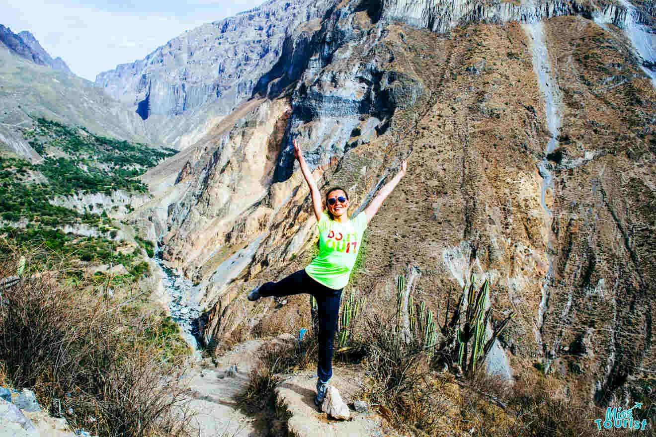 Yulia, la fundadora de este sitio web, vistiendo una camisa verde brillante, se encuentra en equilibrio sobre un camino rocoso frente a un paisaje montañoso, con los brazos y una pierna levantados en una pose alegre, con un cactus a la derecha.