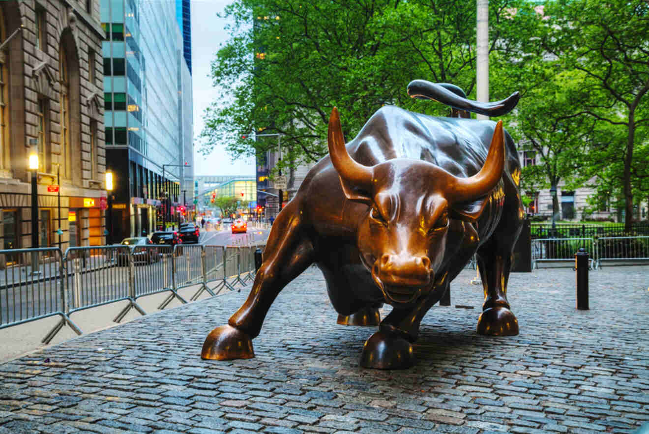 Bronze Charging Bull statue located in the Financial District of New York City, standing on cobblestone pavement, with buildings and trees in the background.