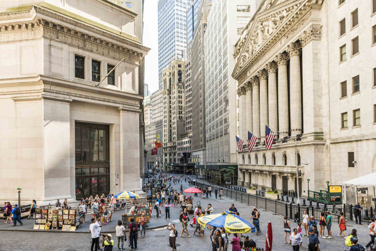 A busy street in a financial district with tall buildings, including a prominent structure adorned with columns and American flags. People are walking and vendors with umbrellas are visible.