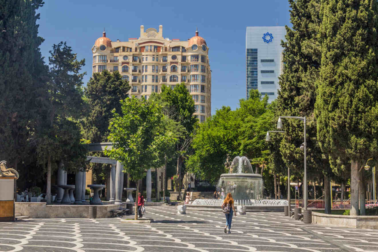 People stroll in a city park with a patterned walkway, surrounded by trees and buildings, including a tall structure with dome-like features and a modern office building in the background.