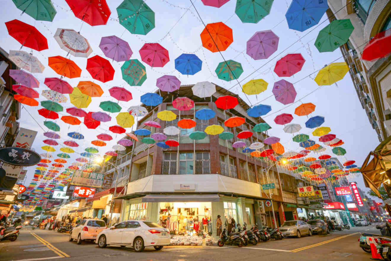 Street scene with numerous colorful umbrellas suspended above, creating a canopy over buildings, shops, and people. Cars and motorbikes are parked along the road.