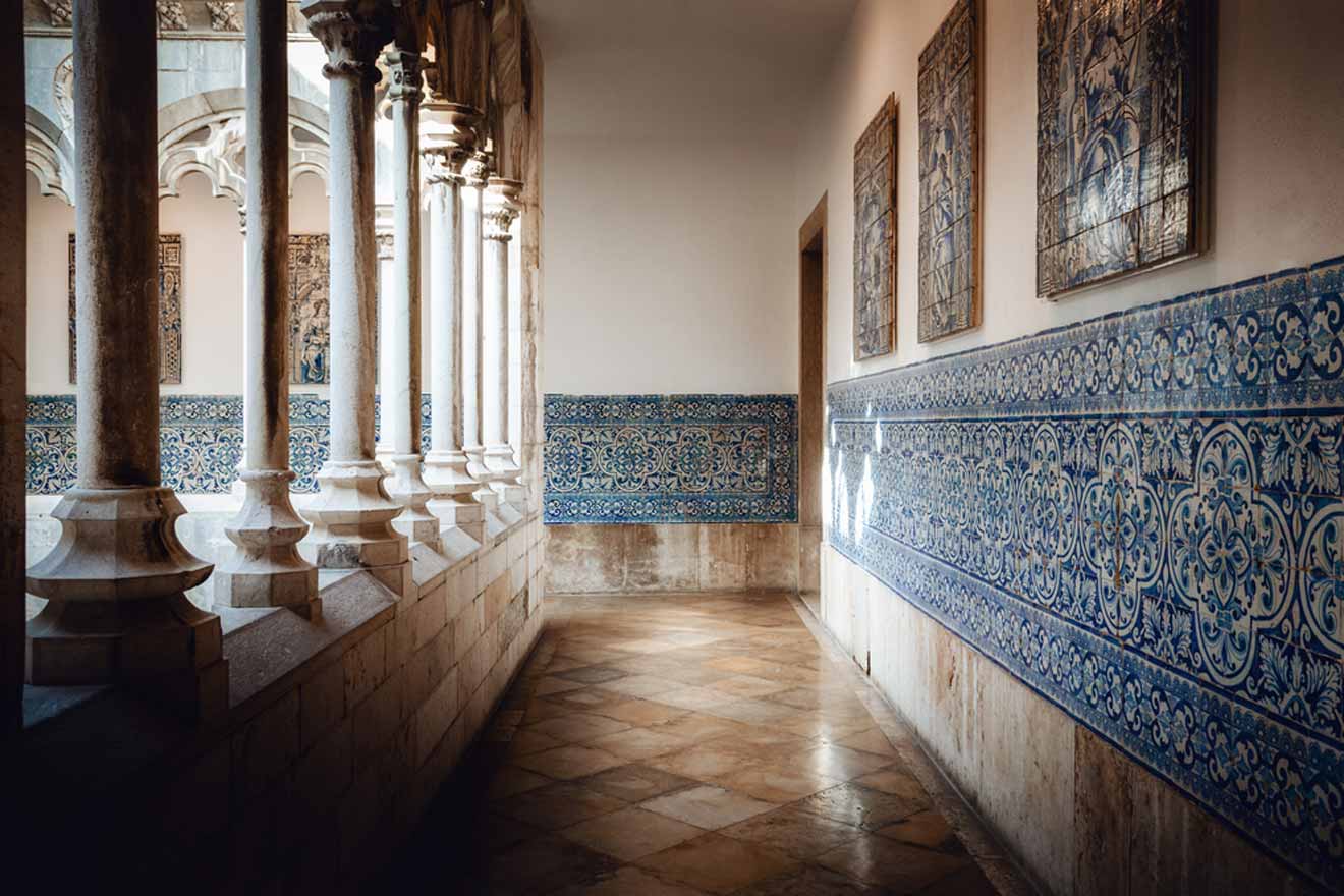 A corridor with ornate blue and white tile work on the lower walls, arched stone columns on the left, and framed artwork hanging on the upper walls. The floor is made of patterned tiles.