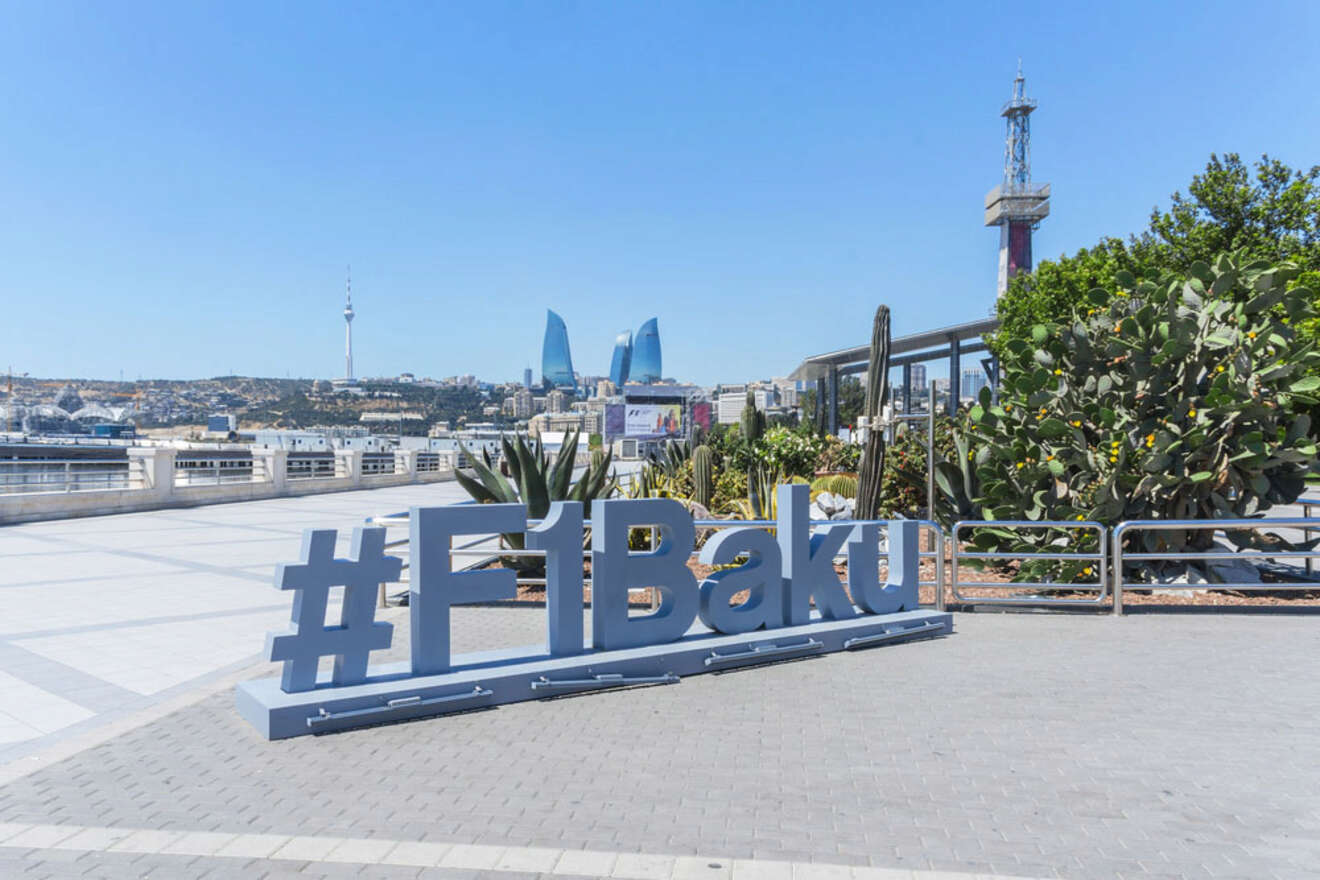 A large #F1Baku sign is displayed on a paved promenade with modern buildings and a television tower visible in the distant cityscape under a clear blue sky.