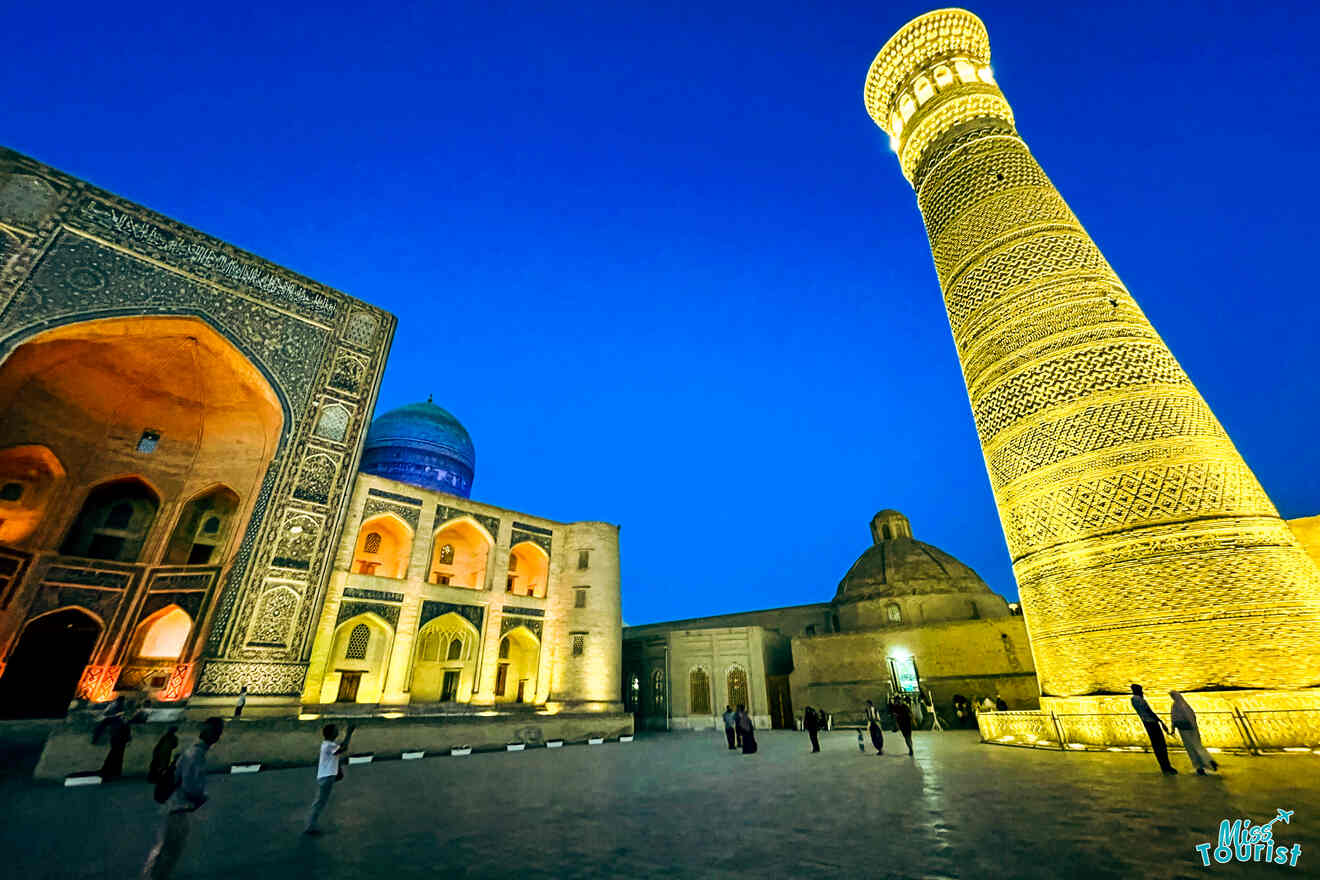 A well-lit, intricately designed mosque and a tall, illuminated minaret against a deep blue evening sky, with a few people walking around the base, highlighting the architectural splendor.