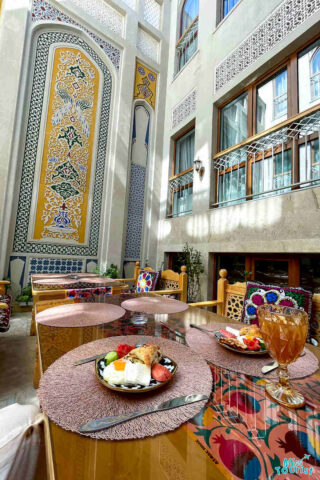 A sunny courtyard with ornate walls, tables set with dishes of food, decorative cushions, and tall windows.