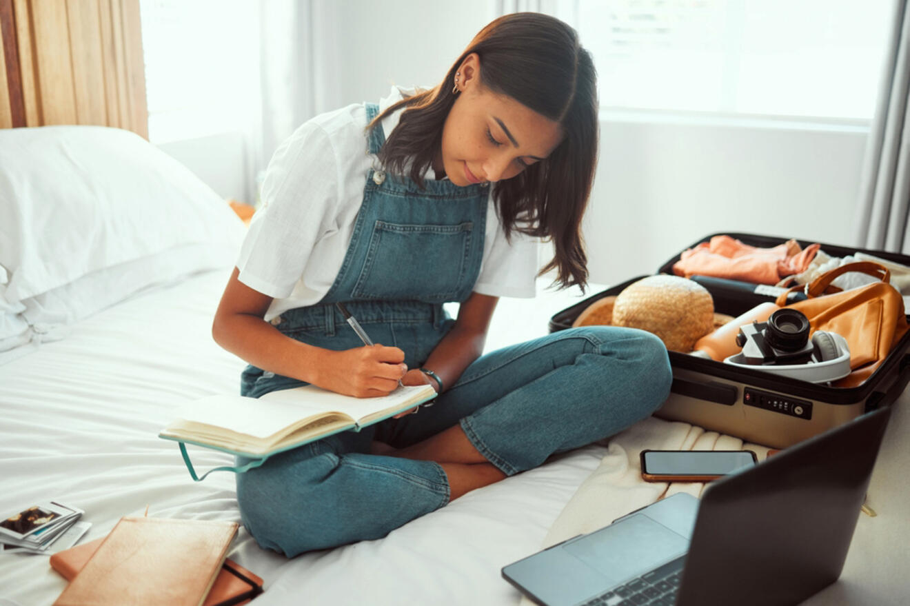 A woman sits on a bed writing in a notebook, with an open suitcase and a laptop beside her.
