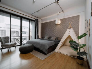 Minimalist bedroom featuring a double bed, teepee tent, floor cushion, chair, potted plant, and large windows with sheer curtains. The room has hardwood floors and a neutral color scheme.