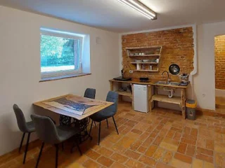 A basement kitchen and dining area featuring a table with four chairs, a kitchenette with a sink and mini fridge, exposed brick walls, tiled floor, and a small window.