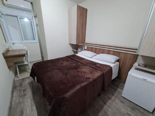 A small bedroom with a double bed covered in a brown blanket, flanked by wooden cabinets, a small desk with a mirror by a window, and a white mini-fridge in the corner.