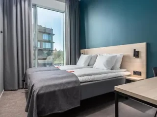 A minimalist bedroom with twin beds, a blue accent wall, a large window, and modern furnishings providing a sleek and comfortable stay.