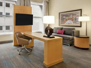 Modern office space with a wooden desk, swivel chair, couch, and two lamps. A large wall-mounted TV and framed artwork can be seen. Floor-to-ceiling windows are in the background.