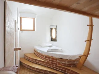 A small bedroom features a unique boat-shaped brick bedframe with a white mattress and folded towels, beneath a small square window and a round porthole-style window.