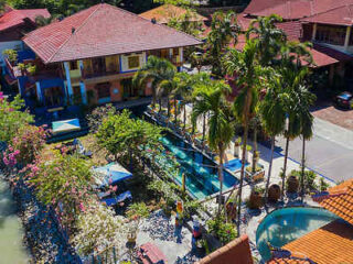 Aerial view of a tropical resort showcasing a central swimming pool, palm trees, lounge chairs, and a surrounding garden with multiple buildings featuring red roofs in the background.
