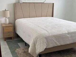 A neatly made bed with a beige upholstered headboard stands next to a wooden bedside table with a lamp on it, in a simple and clean bedroom.