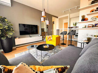 A modern living room with a gray sofa, yellow armchair, wall-mounted TV, and decorative shelving. A potted plant sits in the corner, and a small kitchen area is visible in the background.