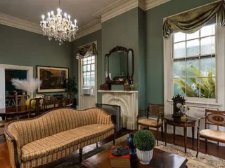 An elegant living room features a striped sofa, a chandelier, vintage furnishings, a fireplace with a mirror above it, and large windows with draped curtains.