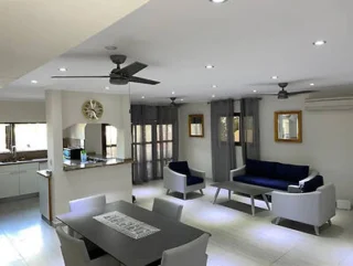 A modern, open-concept living and dining area with gray and white furniture, a ceiling fan, wall clock, and a visible kitchen in the background.