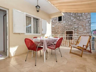 A sunlit patio with a dining table set for four, featuring red chairs and a white tablecloth. There is a wooden building in the background and a striped deck chair to the right.